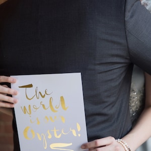 May include: A couple holding a white card with gold lettering that says "The world is our oyster!"