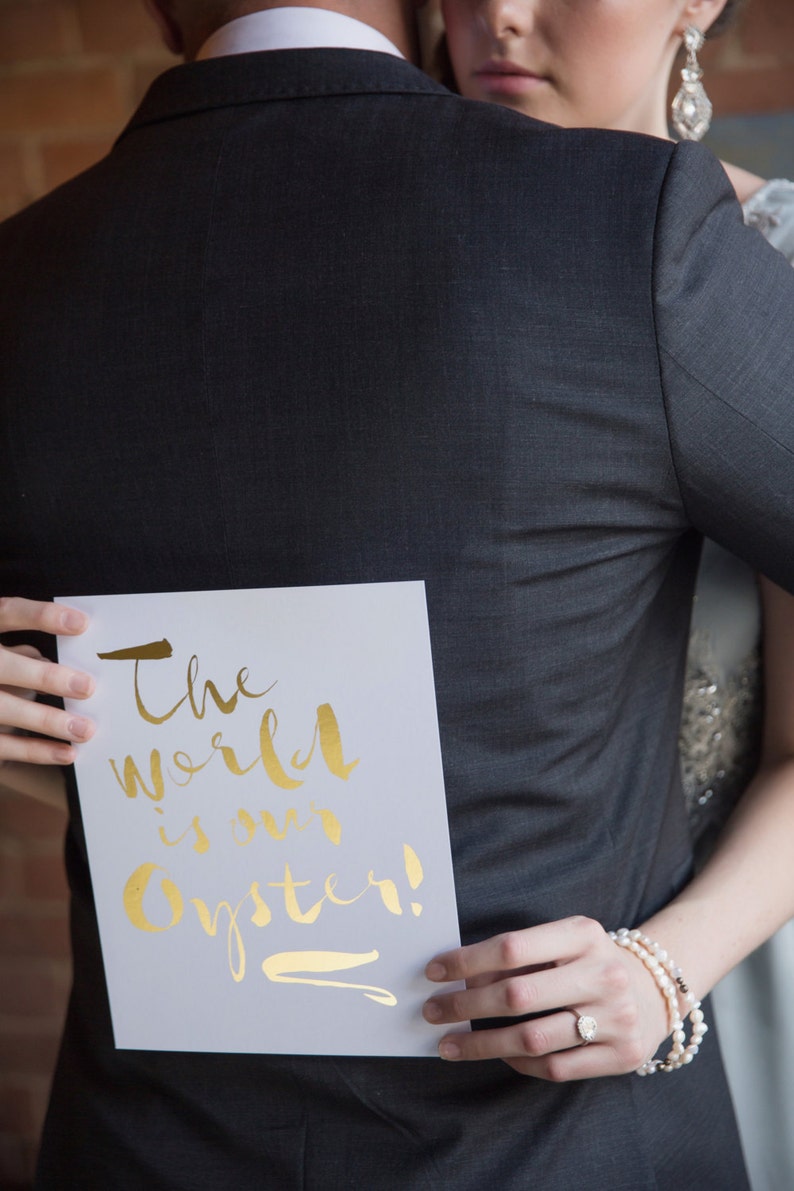 May include: A couple holding a white card with gold lettering that says "The world is our oyster!"