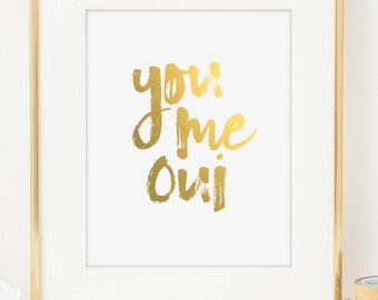May include: A framed print with the words "you me Oui" in gold brush lettering on a white background. The print is in a gold frame and the text is in a handwritten style. The print is from "raincity prints".