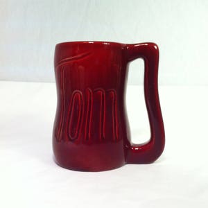 May include: A red ceramic mug with the word "Mom" inscribed on the front. The mug has a unique, curved handle.