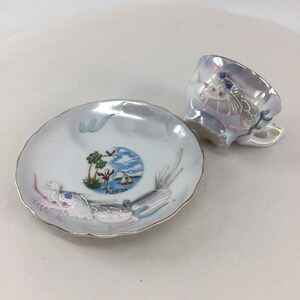 May include: A small, iridescent teacup and saucer set. The white saucer features a Florida scene with palm trees, a sailboat, and the word "Florida." The cup and saucer have gold trim and decorative elements.