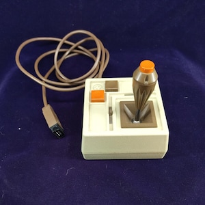 May include: A beige video game joystick controller featuring a brown tapered handle with an orange top button, a square orange button, a small brown square button, and a long brown cord ending in a black multi-pin connector plug.