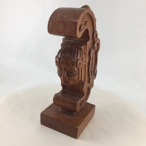 May include: A wooden sculpture of a stylized human face with intricate details. The sculpture is carved from a single piece of wood and features a raised base.