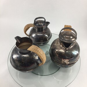 May include: A silver tea set with a glass serving tray. The teapot and creamer have a round, bulbous shape with a woven wicker handle. The sugar bowl has a lid with a woven wicker handle.