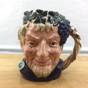 Puede incluir: Una jarra de cerámica con forma de cabeza de hombre con una corona de hojas y uvas. La jarra tiene un asa marrón y blanca y una túnica azul y morada.