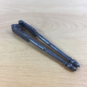 May include: A vintage metal nutcracker with a simple design. The nutcracker is made of iron and has a dark gray finish. It has two long handles with rounded ends.