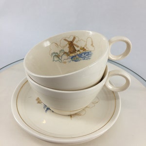 May include: A set of two white teacups and a saucer with a gold rim. The cups and saucer feature a delicate design of a windmill and clouds in shades of blue and brown.