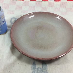 May include: A large, round, light gray ceramic plate with a reddish-brown rim. The plate is slightly textured and has a subtle sheen.