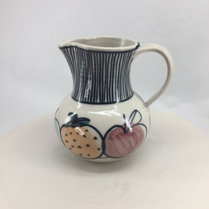 May include: A white ceramic pitcher with a black and white striped design around the top. The pitcher has a handle and is decorated with hand-painted fruit, including a red apple, a yellow orange, and a green pear.