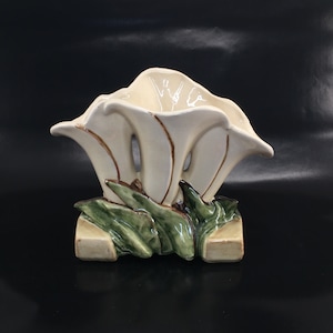 May include: A white ceramic flower vase with green leaves and brown accents. The vase is shaped like a flower with three petals.