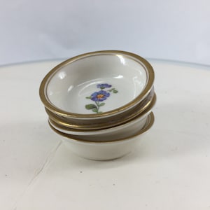 May include: A stack of four small white ceramic bowls with a gold rim and a blue flower design.