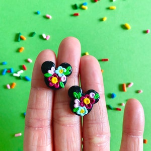 May include: A pair of black stud earrings with a floral design. The flowers are pink, yellow, white, and red. The earrings are shaped like Mickey Mouse ears.