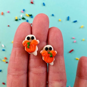 May include: A pair of white ghost earrings with orange pumpkin faces. Each ghost is holding a small green pumpkin stem in its mouth.