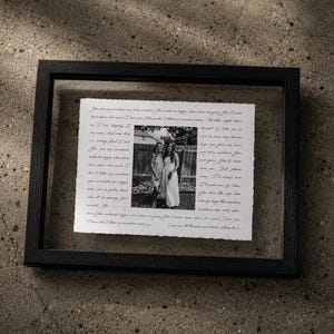 May include: A black rectangular floating frame containing a white piece of paper with deckled edges. The paper features a central black and white photograph of two people standing outdoors, surrounded by handwritten cursive text in black ink.