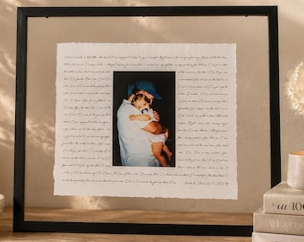 May include: A black rectangular floating frame containing a white piece of paper with deckled edges. The paper features a central color photograph of a person holding a baby, surrounded by handwritten cursive text in black ink on both sides.