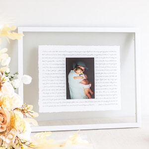 May include: A white framed photo print featuring a photograph of a person holding a child. The photo is centered on white paper with handwritten text around it. The frame is white, and the background is white.