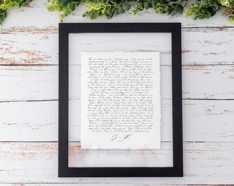 May include: A black rectangular floating frame containing a white paper with deckled edges featuring handwritten black cursive lyrics. The text includes the initials J + K at the bottom. The frame is displayed against a white wood background.