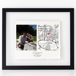 May include: Framed print featuring a photograph of a couple and a map with a red heart marking a location. The print is labeled "Engaged" with names and a date. The frame is black.