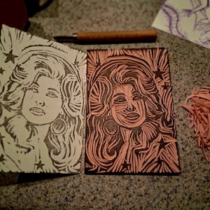 May include: A rectangular pink linoleum block carved with a portrait of a person with voluminous hair and hoop earrings, surrounded by stars. Beside it is a black ink print of the same design on white paper, with wood carving tools.