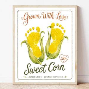 May include: A framed print featuring the text Grown With Love at the top and Sweet Corn at the bottom. Two yellow baby footprints are positioned inside green corn husks. A small heart icon displays 50 cents. Text reads LOCALLY GROWN LOVINGLY HARVESTED.