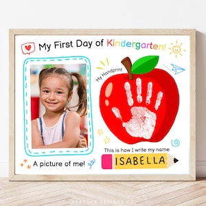 May include: A framed print for a child's first day of kindergarten. The design includes a photo frame, a red apple with a handprint, and the text "My First Day of Kindergarten!" and "This is how I write my name ISABELLA."