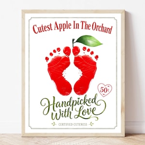 May include: A framed print featuring red baby footprints shaped like an apple with a green leaf. The text reads Cutest Apple In The Orchard, Handpicked With Love, and Certified Cuteness, with a small heart icon containing the text 50 cents.