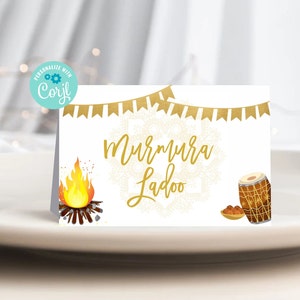 May include: A white card with gold text and illustrations. The text reads "Murmura Ladoo" in a script font. Illustrations include a bonfire, a drum, and a small bowl of food. A banner of gold flags is at the top.