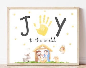 May include: A framed print with a light wood frame. The print features the words "JOY to the world" with a yellow handprint in place of the "O". Below is a watercolor illustration of the nativity scene with stars.