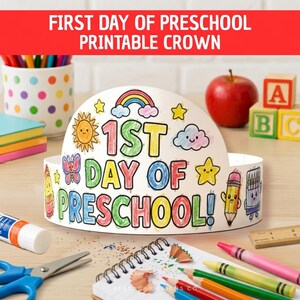 Op de afbeelding: Een witte papieren kroon met de kleurrijke tekst 1ST DAY OF PRESCHOOL!, versierd met illustraties van een regenboog, zon, wolk, vlinder, sterren, potlood en krijtje. De kroon staat op een oppervlak met diverse schoolbenodigdheden.