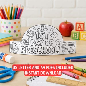 Op de afbeelding: Een witte papieren kroon met zwarte lijntekeningen met de tekst 1ST DAY OF PRESCHOOL!, omringd door illustraties van een zon, regenboog, wolken, sterren, een vlinder, een rugzak, een potlood en een doosje kleurpotloden voor activiteiten.