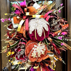 May include: A decorative wreath featuring two velvet pumpkins in deep burgundy and burnt orange, accented with gold glitter, layered ribbons in plaid and solid jewel tones, dried foliage, and small purple flowers, hanging on a dark door.