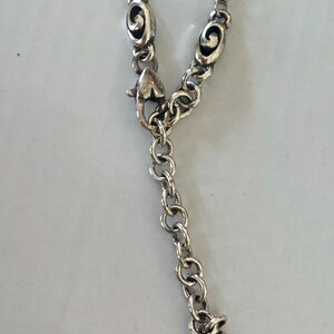 May include: Silver-toned necklace with a heart-shaped charm. The necklace features a series of small, round links and decorative oval-shaped elements. The heart charm has a cursive "B" engraved on it. The clasp is a lobster claw style.
