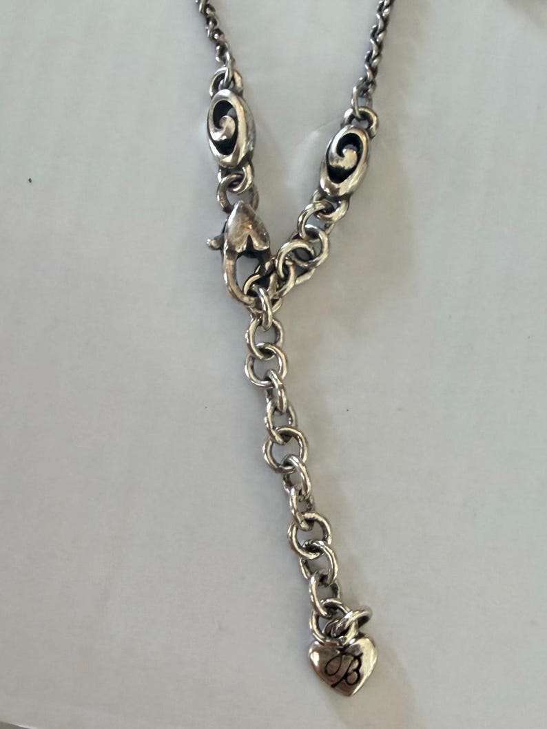 May include: Silver-toned necklace with a heart-shaped charm. The necklace features a series of small, round links and decorative oval-shaped elements. The heart charm has a cursive "B" engraved on it. The clasp is a lobster claw style.