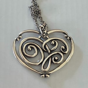 May include: Silver-toned pendant necklace featuring a heart-shaped design with intricate scrollwork. The pendant hangs from a delicate chain. The design includes swirling patterns and a decorative bail.