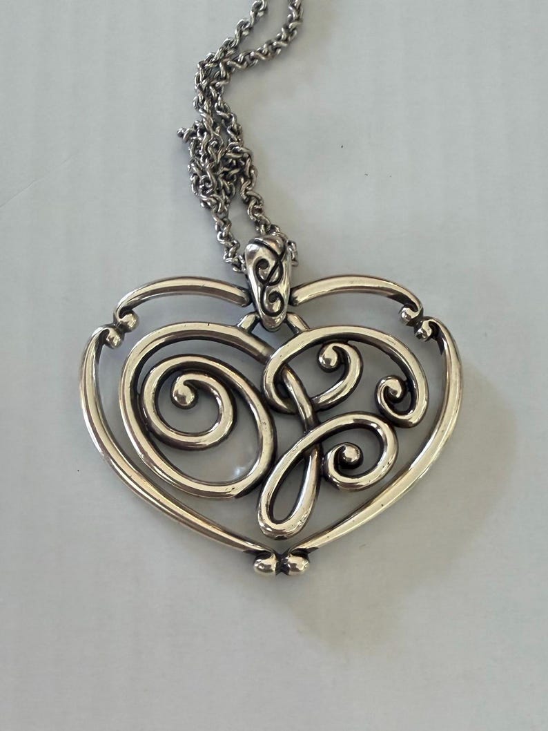 May include: Silver-toned pendant necklace featuring a heart-shaped design with intricate scrollwork. The pendant hangs from a delicate chain. The design includes swirling patterns and a decorative bail.