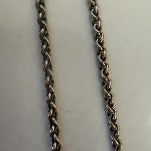 May include: Close-up of two silver-colored chains with a braided rope design. The chains are positioned vertically against a light-colored background. The links are tightly woven, creating a textured surface.