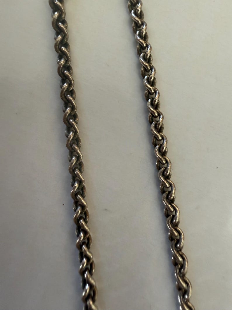 May include: Close-up of two silver-colored chains with a braided rope design. The chains are positioned vertically against a light-colored background. The links are tightly woven, creating a textured surface.