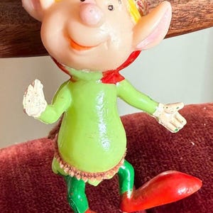 May include: A vintage Christmas elf ornament. The elf has a green body, red hat and shoes, and a smiling face. The elf's hat has a white pom-pom. The elf is wearing a red collar and has white gloves.
