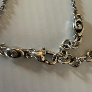 May include: Close-up of a silver-toned necklace clasp and chain. The clasp is heart-shaped, and the chain links are small and circular. The necklace has a vintage or antique appearance.