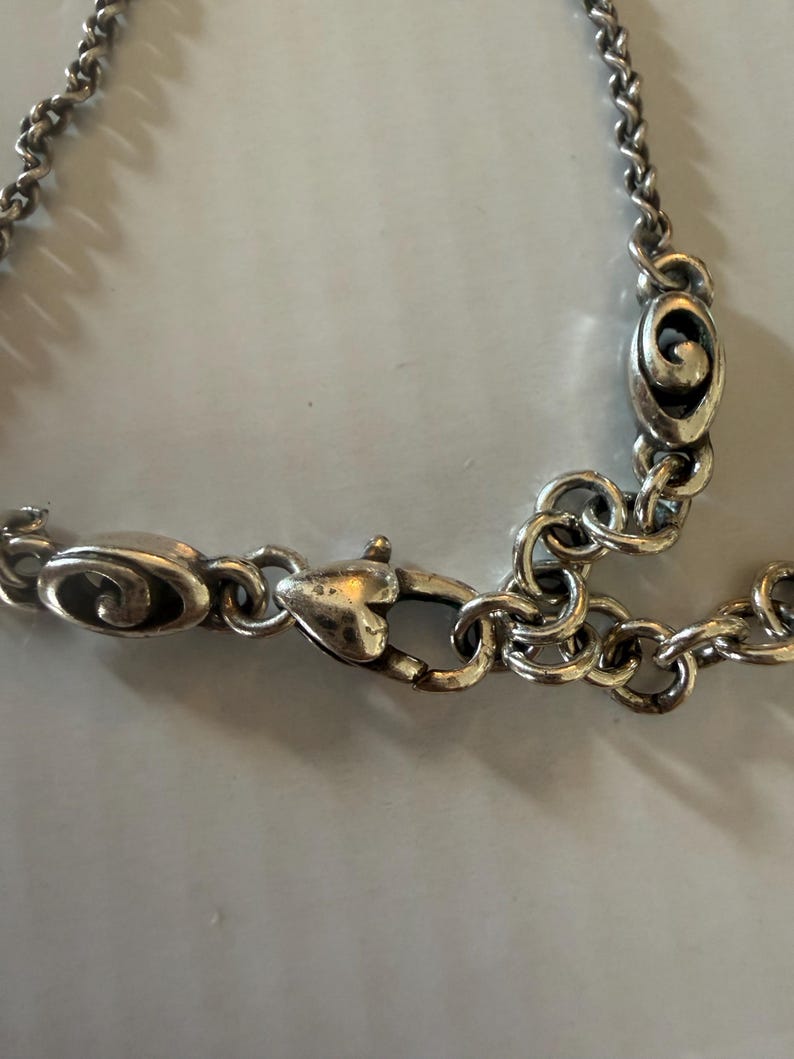 May include: Close-up of a silver-toned necklace clasp and chain. The clasp is heart-shaped, and the chain links are small and circular. The necklace has a vintage or antique appearance.