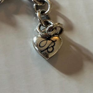 May include: Close-up of a silver-toned charm bracelet. The bracelet features a heart-shaped charm with the letter "B" engraved on it. The charm is attached to a chain link bracelet.