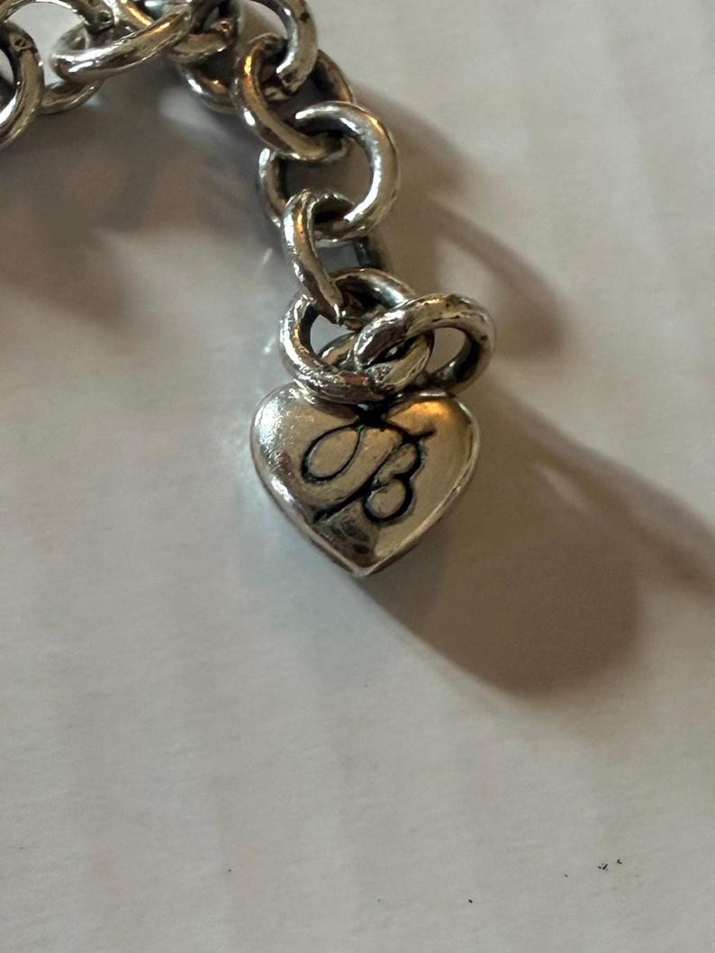 May include: Close-up of a silver-toned charm bracelet. The bracelet features a heart-shaped charm with the letter "B" engraved on it. The charm is attached to a chain link bracelet.