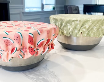 May include: Two reusable bowl covers, one with a watermelon print and the other with a green floral print. The covers are made of fabric and are sitting on a white countertop.