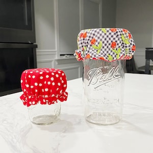 Op de afbeelding: Twee glazen potten met stoffen deksels. De grotere pot heeft een zwart-wit geruit deksel met rode kersen en groene bladeren. De kleinere pot heeft een rood gestippeld deksel. Beide potten zijn van helder glas en hebben het woord "Ball" erop gedrukt.