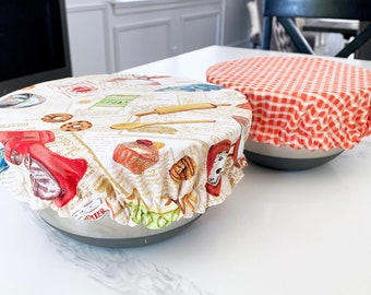 May include: Two reusable food covers on bowls. One cover is red and white gingham, the other is white with a colorful vintage print of food and kitchen items.