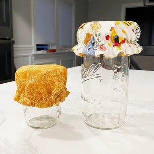 Può includere: Due barattoli di vetro con coperchi in tessuto. Il barattolo più grande ha un coperchio in tessuto bianco con un motivo floreale e un pollo. Il barattolo più piccolo ha un coperchio in tessuto giallo con un motivo floreale.