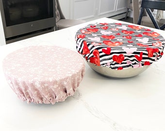 May include: Two reusable bowl covers, one pink with a floral pattern and one black and white with red hearts. The covers are made of fabric and are sitting on a white countertop.