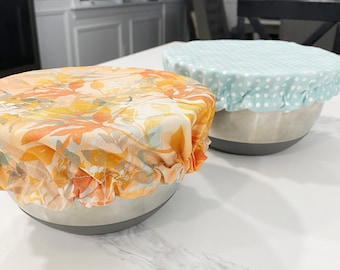 May include: Two bowls covered with fabric bowl covers. One bowl cover is a floral pattern in shades of orange, yellow, and white. The other bowl cover is a light blue with white polka dots.