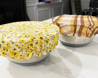 May include: Two reusable bowl covers, one with a yellow floral pattern and the other with a brown and yellow plaid pattern.
