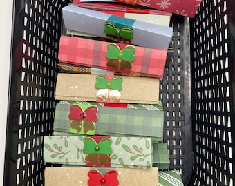 May include: A black basket filled with rectangular gift boxes. The boxes are wrapped in various festive papers, including red with snowflakes, plaid, and green with white sprigs. Each box is adorned with a decorative bow in red, green, and gold.